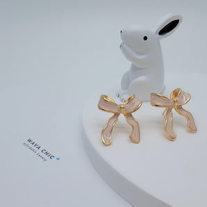 Close-up of the Bubblegum Bow Studs featuring delicate titanium steel bows coated in glossy pink enamel with gold-plated accents. The enamel's smooth finish and the detailed bow shape reflect light softly, highlighting the sweet, whimsical design.

