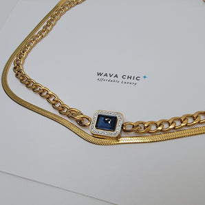 Close-up of Sapphire Reign necklace showing the intricate contrast of a flat herringbone chain and a thick curb chain, with a royal blue imitation sapphire pendant encircled by a diamond-like halo.
