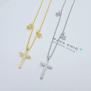 Close-up of the Mini Grace Necklace showcasing the polished sterling silver cross pendant, with a delicate CZ heart charm and a tiny star charm dangling asymmetrically on either side. The craftsmanship highlights shine, detail, and delicate proportions.